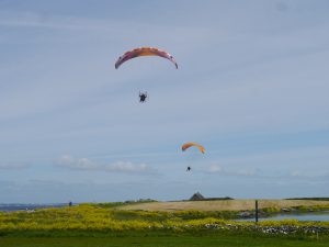 yellow paraglider and orange & white paraglider flying over yellow mowed fields