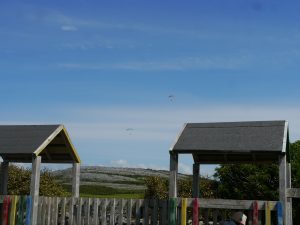 Paragliders in the distance. Wooden playground structure in foreground. Grey burren mountain in background.