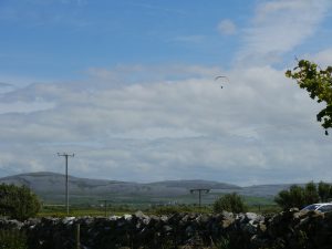 Paraglider in the distance. Burren wall (grey stones) in foreground. Grey burren mountains in background.