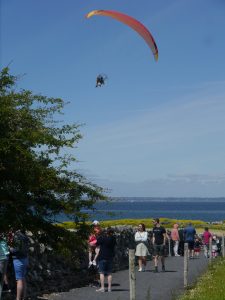 Orange paraglider against a blue sky. People walking on path beneeth him. Deep blue sean in the background.